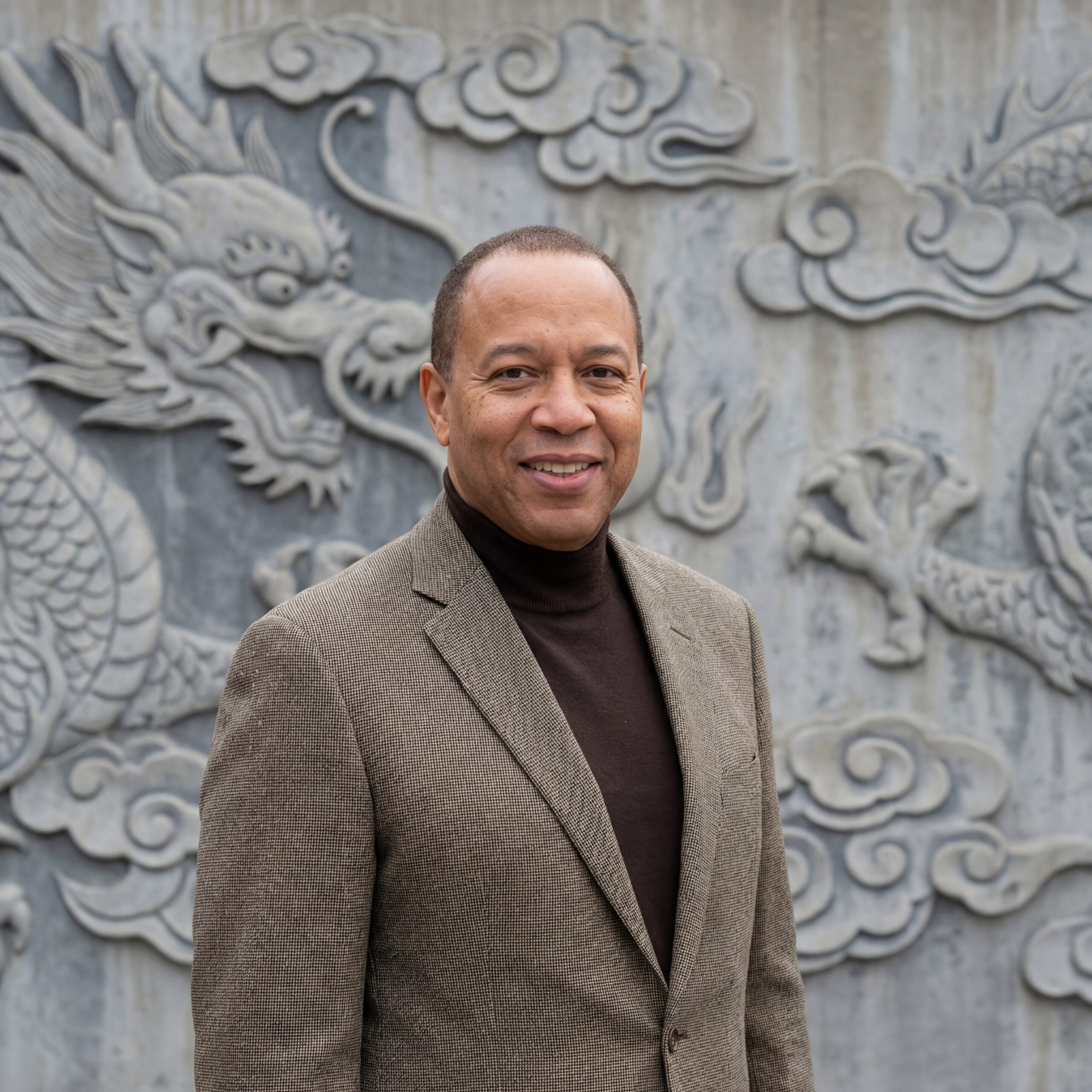 Light Brown Skinned Man Wearing A suit - Enno Momentum Light Brown skinned man wearing suit with a turtleneck smiling with a gray dragon background wall