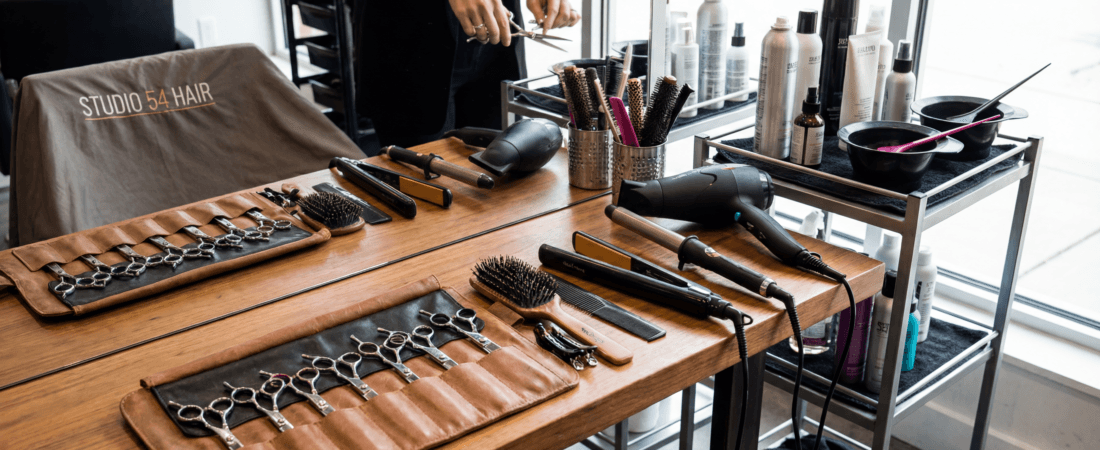 Hair-styling station with a row of scissors in a brown leather roll, brushes, and a hairdryer on a wooden table.