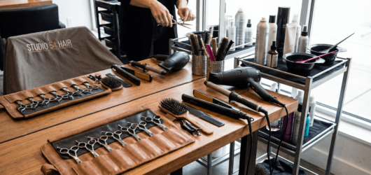 Hair-styling station with a row of scissors in a brown leather roll, brushes, and a hairdryer on a wooden table.